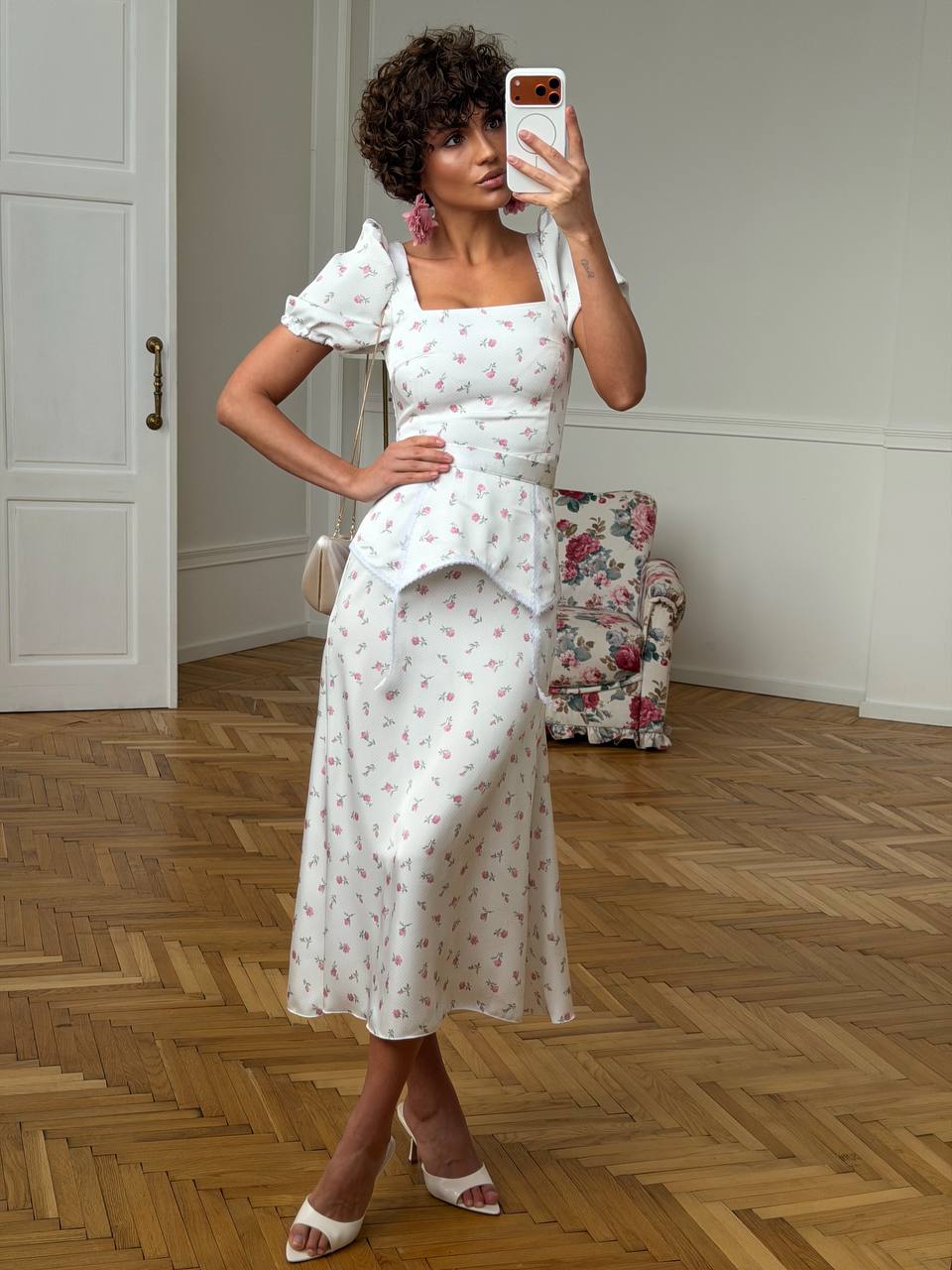Woman taking a mirror selfie wearing a white floral dress in a room with wooden flooring.