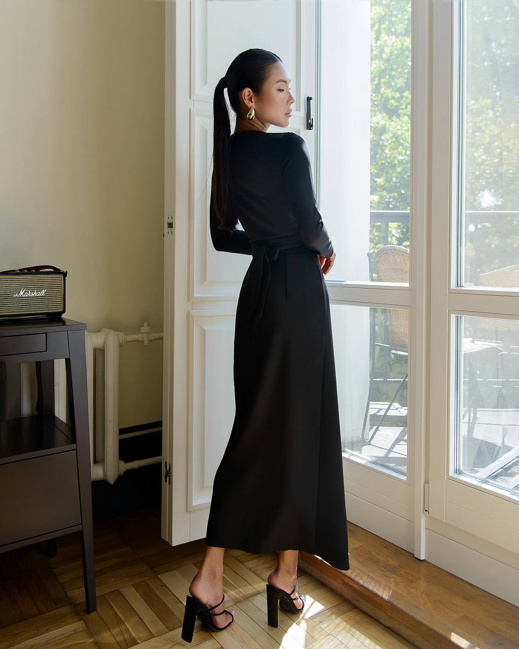 Woman in a black dress standing by a window in a well-lit room.