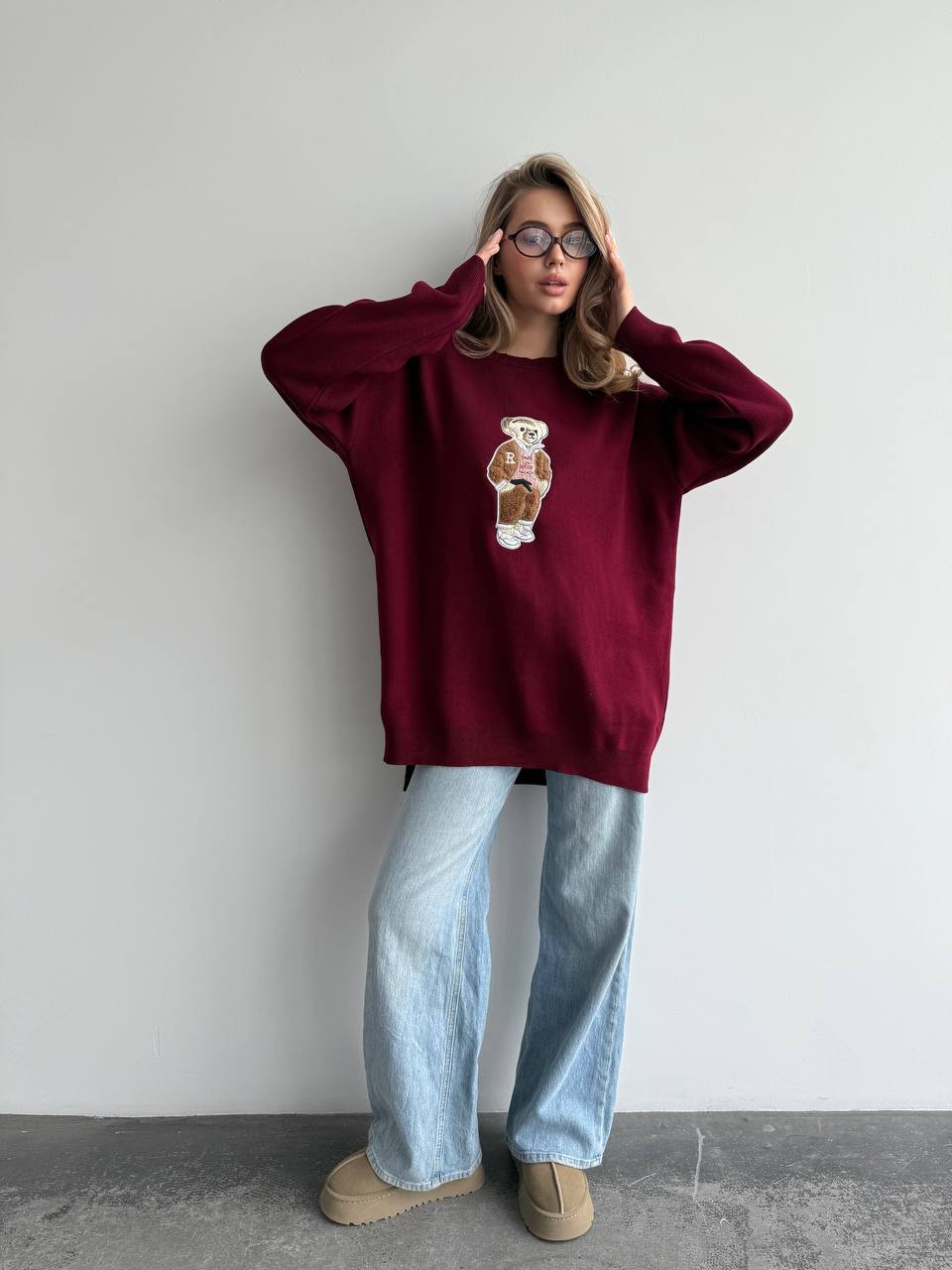 Person wearing a burgundy sweatshirt with a graphic design and light blue jeans against a plain background