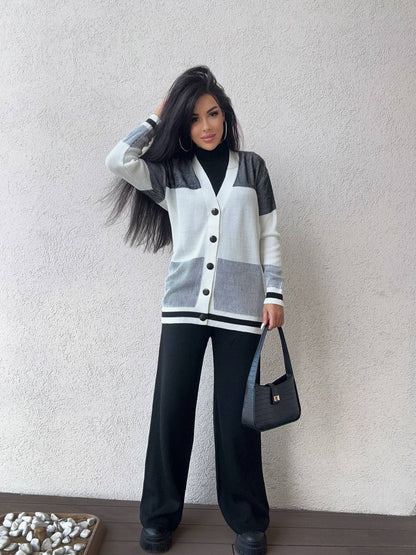 Woman wearing a gray and black cardigan with black pants against a plain wall.