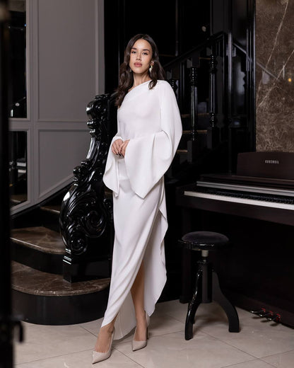 Woman in a white dress standing in a room with a piano and decorative chair.