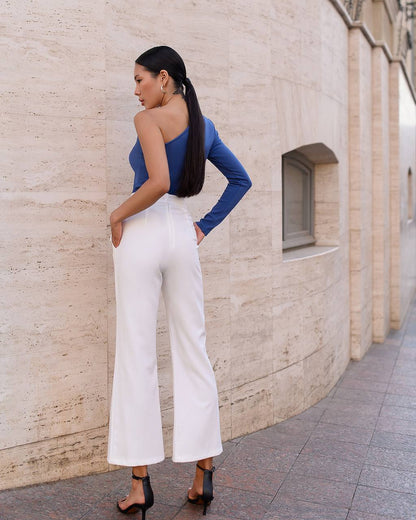 Woman in a blue top and white pants standing against a beige wall.