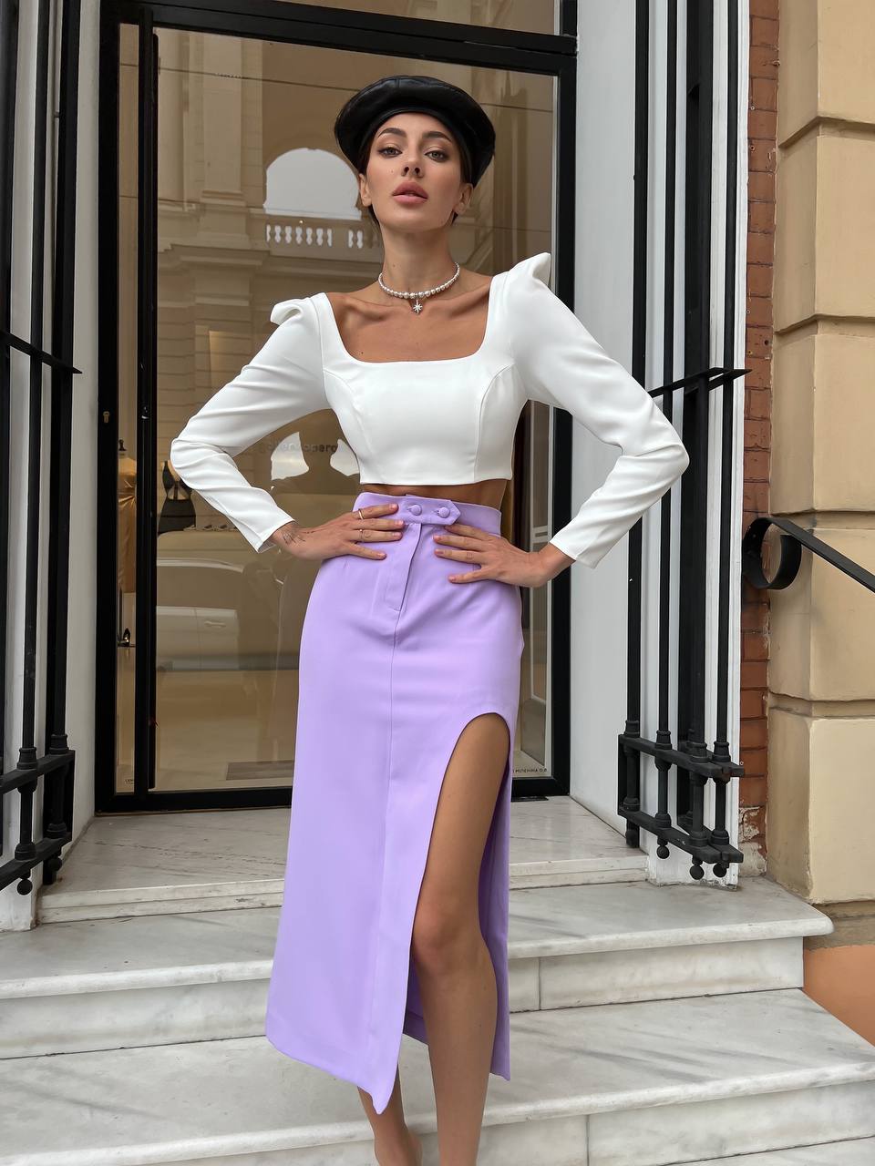 Woman in a white top and purple skirt standing in front of a building entrance.