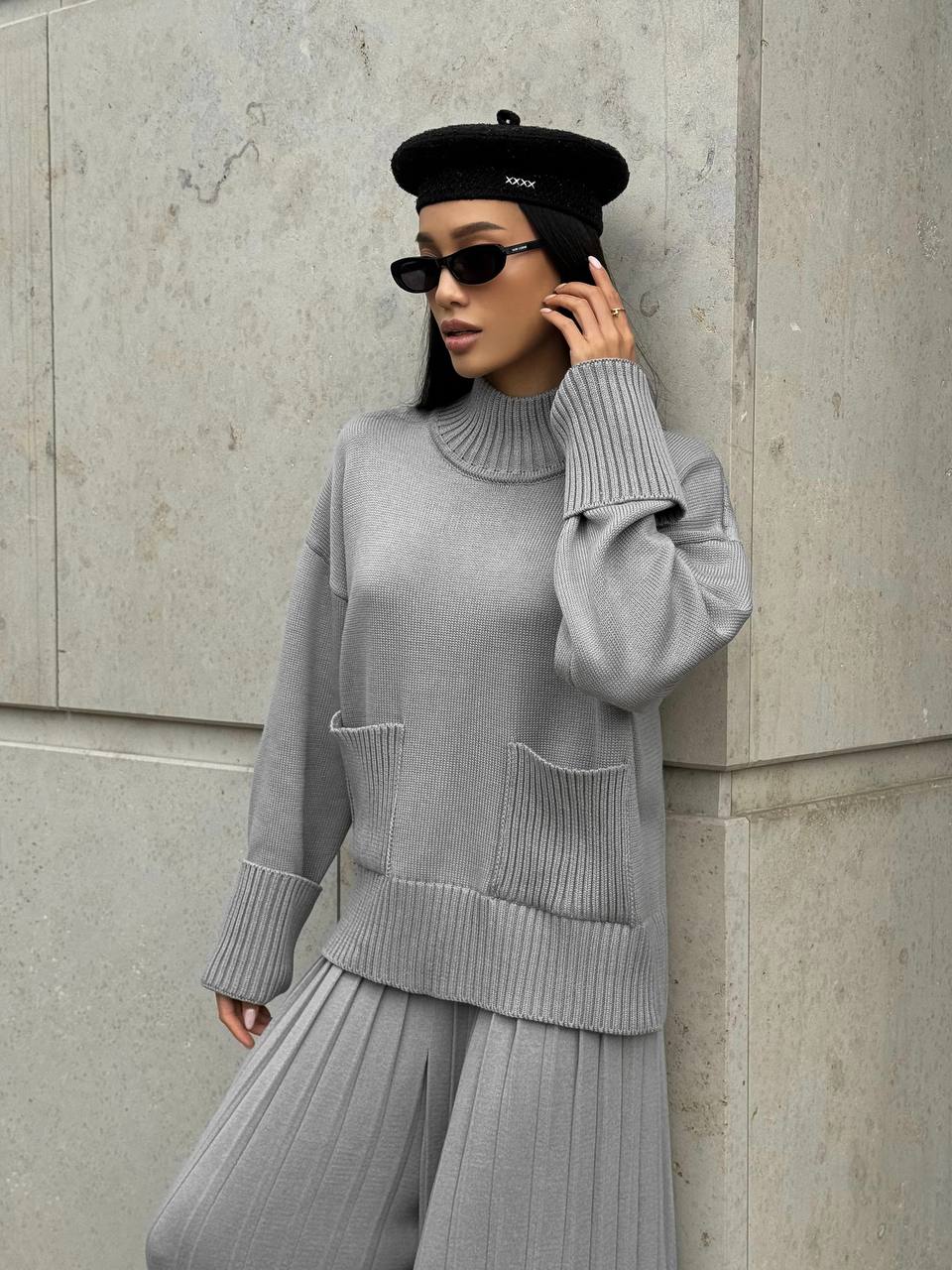 Person wearing a gray sweater and pants leaning against a concrete wall.