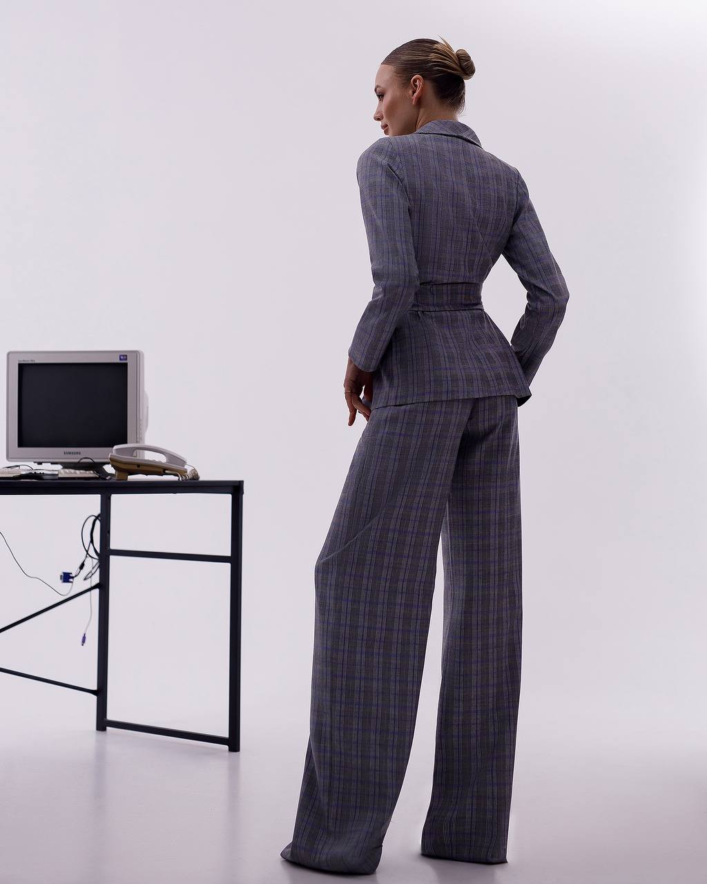 Person wearing a gray checkered suit standing in front of a white wall with a computer desk.