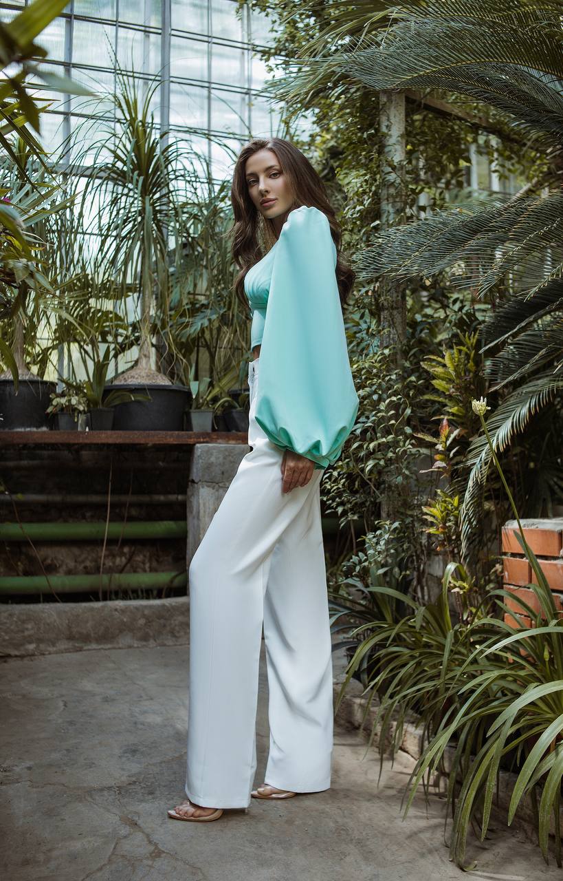 Woman in a green top and white pants standing in a greenhouse filled with plants.