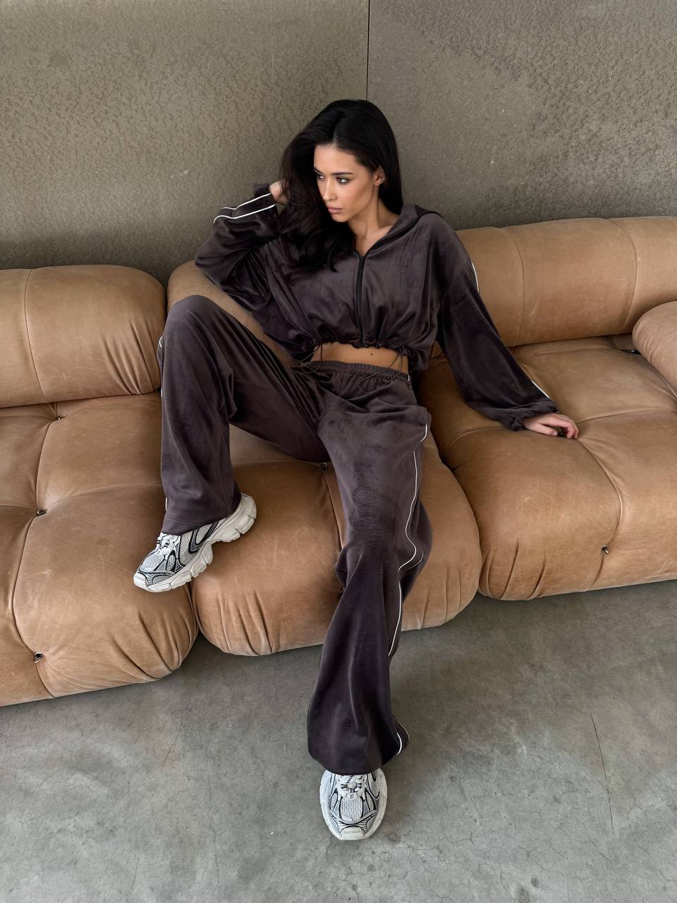 Woman in a brown tracksuit sitting on a beige sofa against a gray wall.