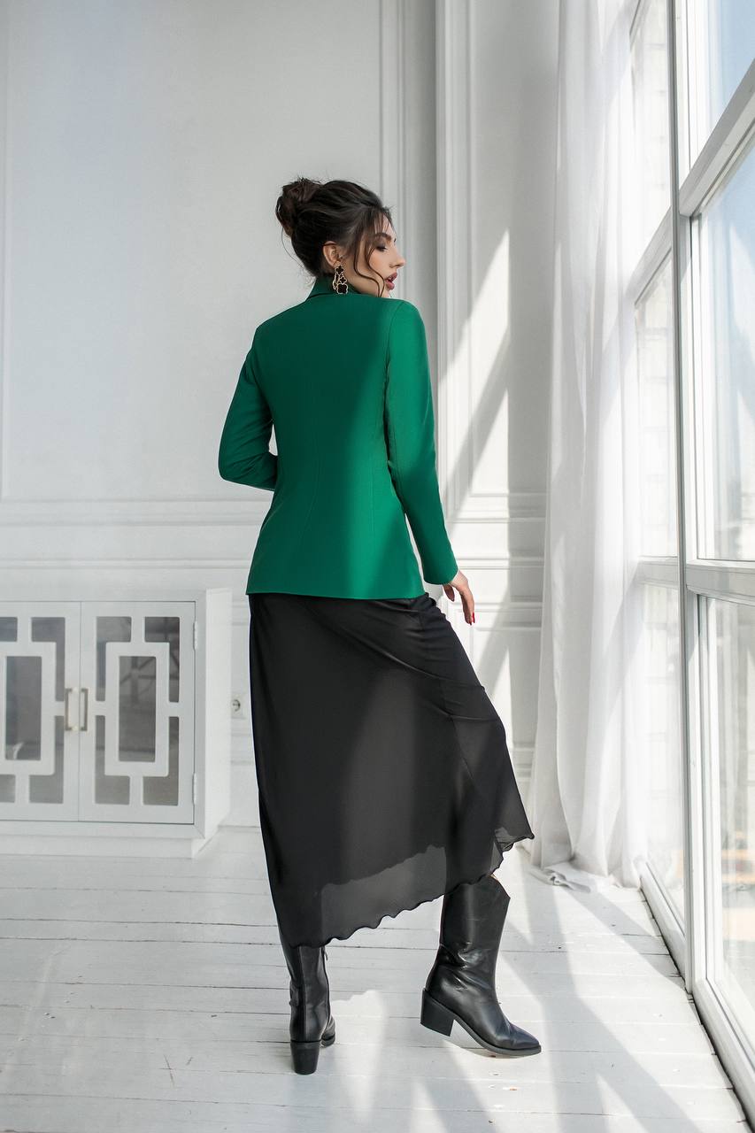 Woman in a green blazer and black skirt standing in a bright room with large windows.