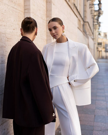 Two models standing on a city street. One is wearing a structured white suit with a matching top, and the other is wearing a dark brown blazer. They are posed against a light-colored stone building wall.