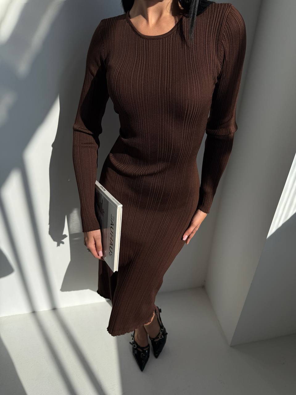 Woman wearing a brown ribbed dress holding a book against a white wall.