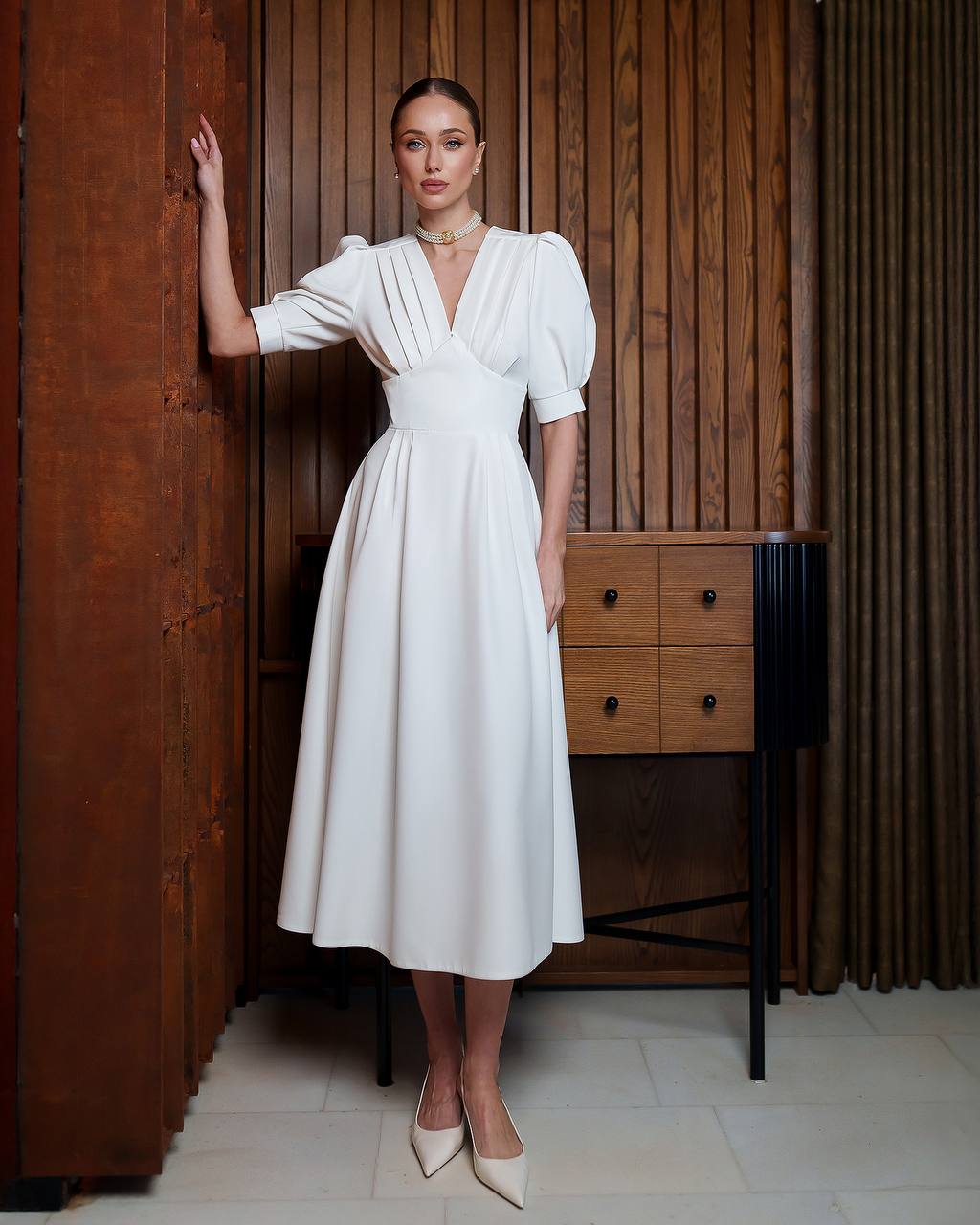 Woman in a white dress standing in a room with wooden paneling and furniture.