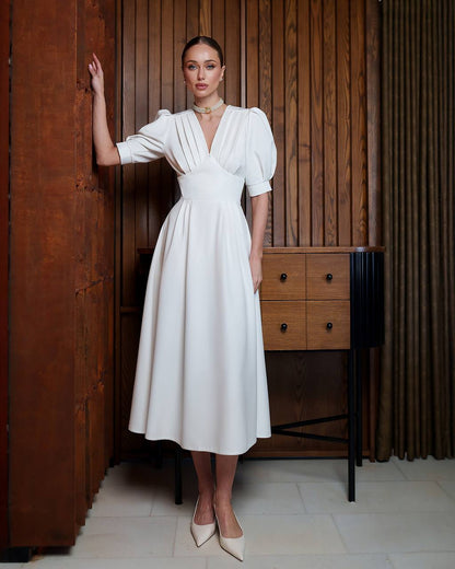 Woman in a white dress standing in a room with wooden paneling and furniture.