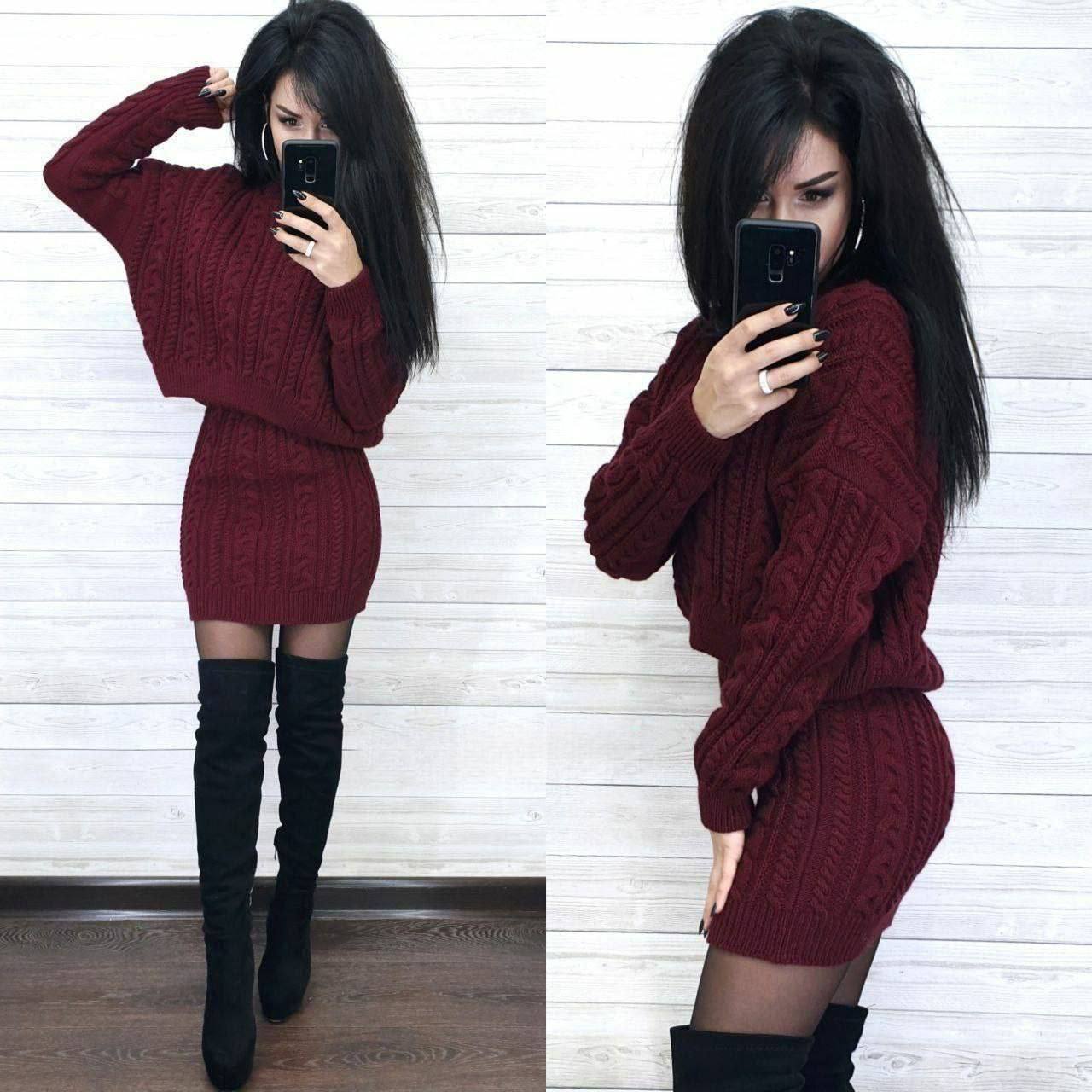 Woman wearing a burgundy cable knit sweater set with black knee-high boots against a white wooden panel background.