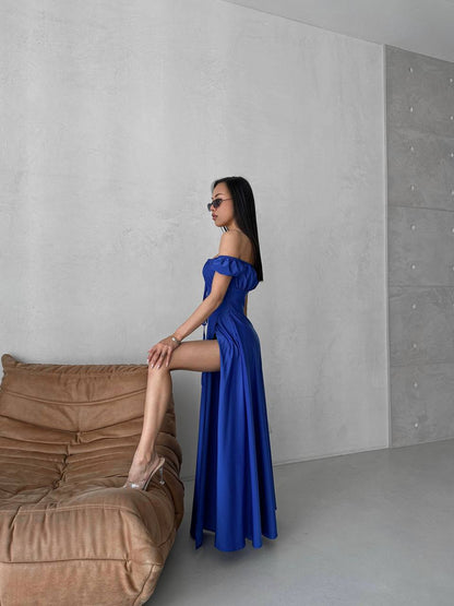 Woman in a blue dress posing next to a brown sofa against a gray wall.