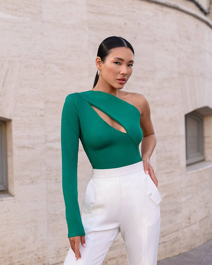 Woman wearing a green one-shoulder top with cut-out design and white pants against a beige wall.