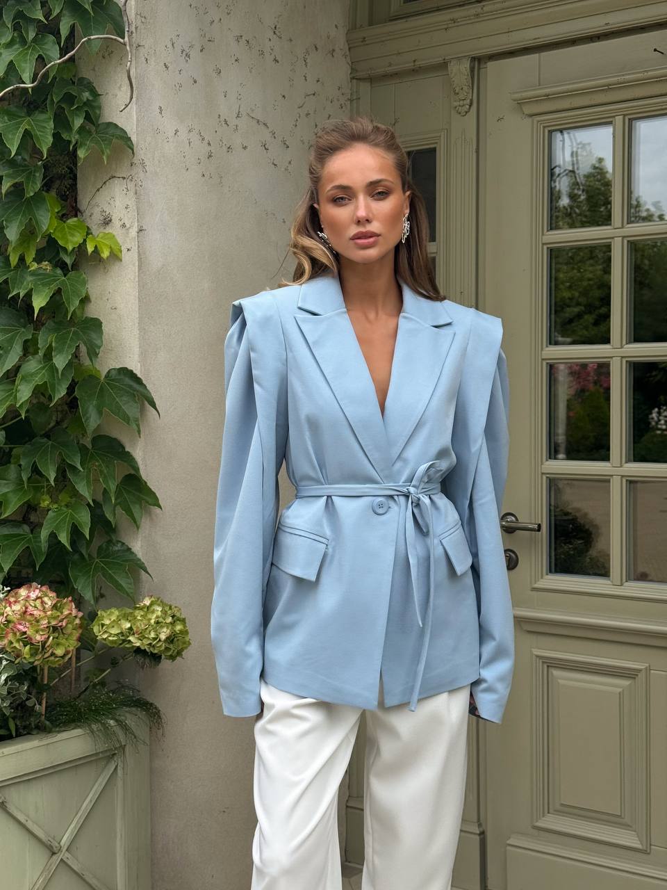 Woman wearing a light blue blazer and white pants standing in front of a door with greenery.