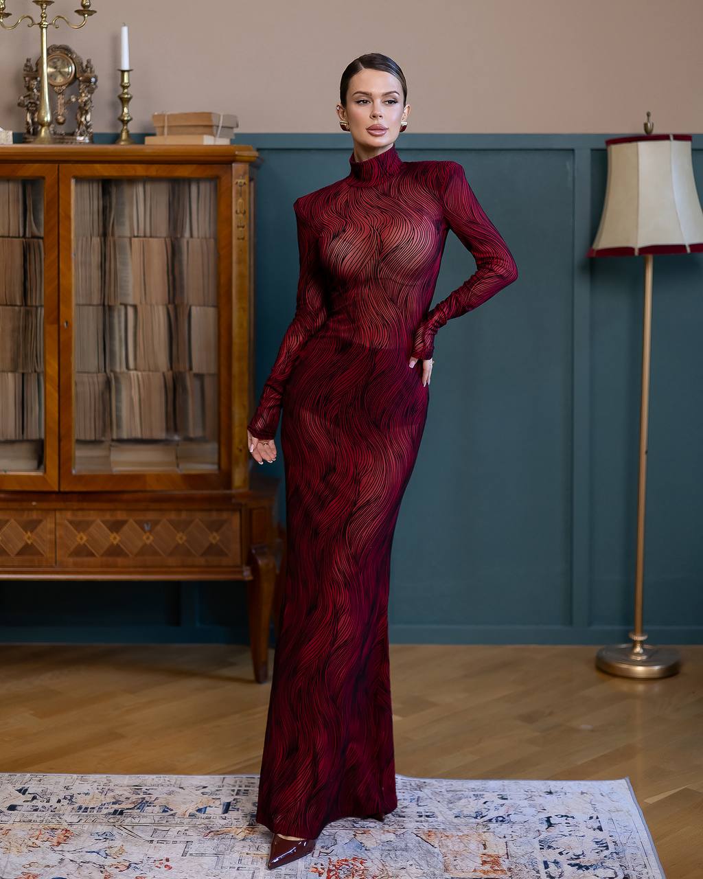 Woman in a burgundy evening gown standing in a room with wooden furniture and a lamp.