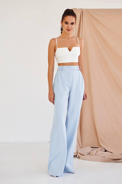 Woman wearing a white top and light blue pants against a beige curtain.