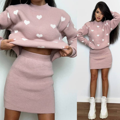 Woman wearing a pink sweater and skirt set with heart patterns, standing against a white wall.