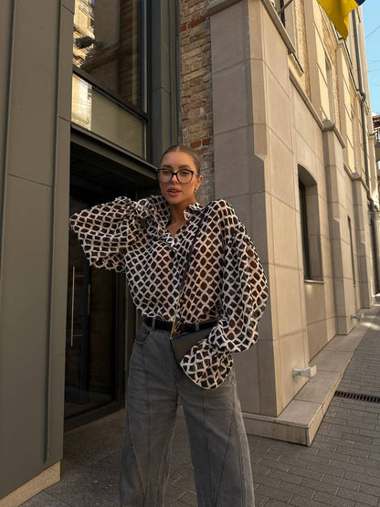 Person wearing a patterned shirt and jeans standing on a street with buildings in the background