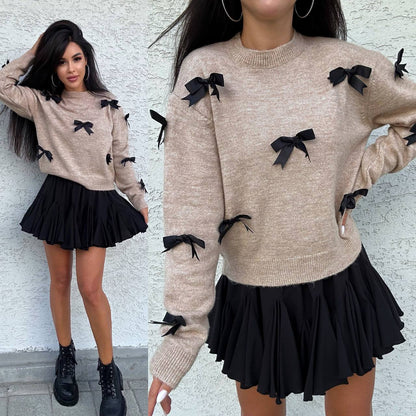 Woman wearing a beige sweater with black bows and a black skirt against a white wall.