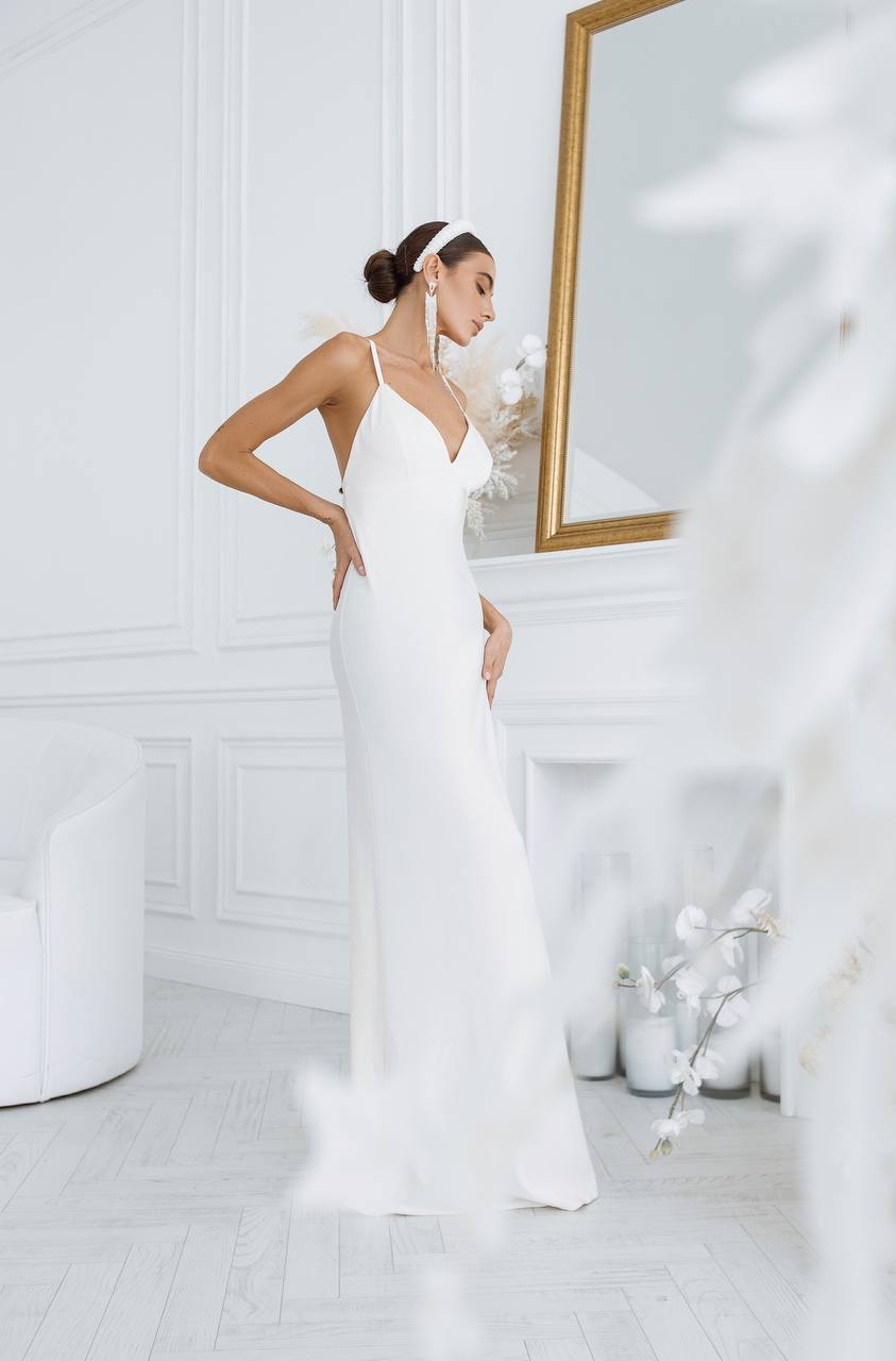Woman in a white dress standing in a bright, minimalistic room with a mirror.