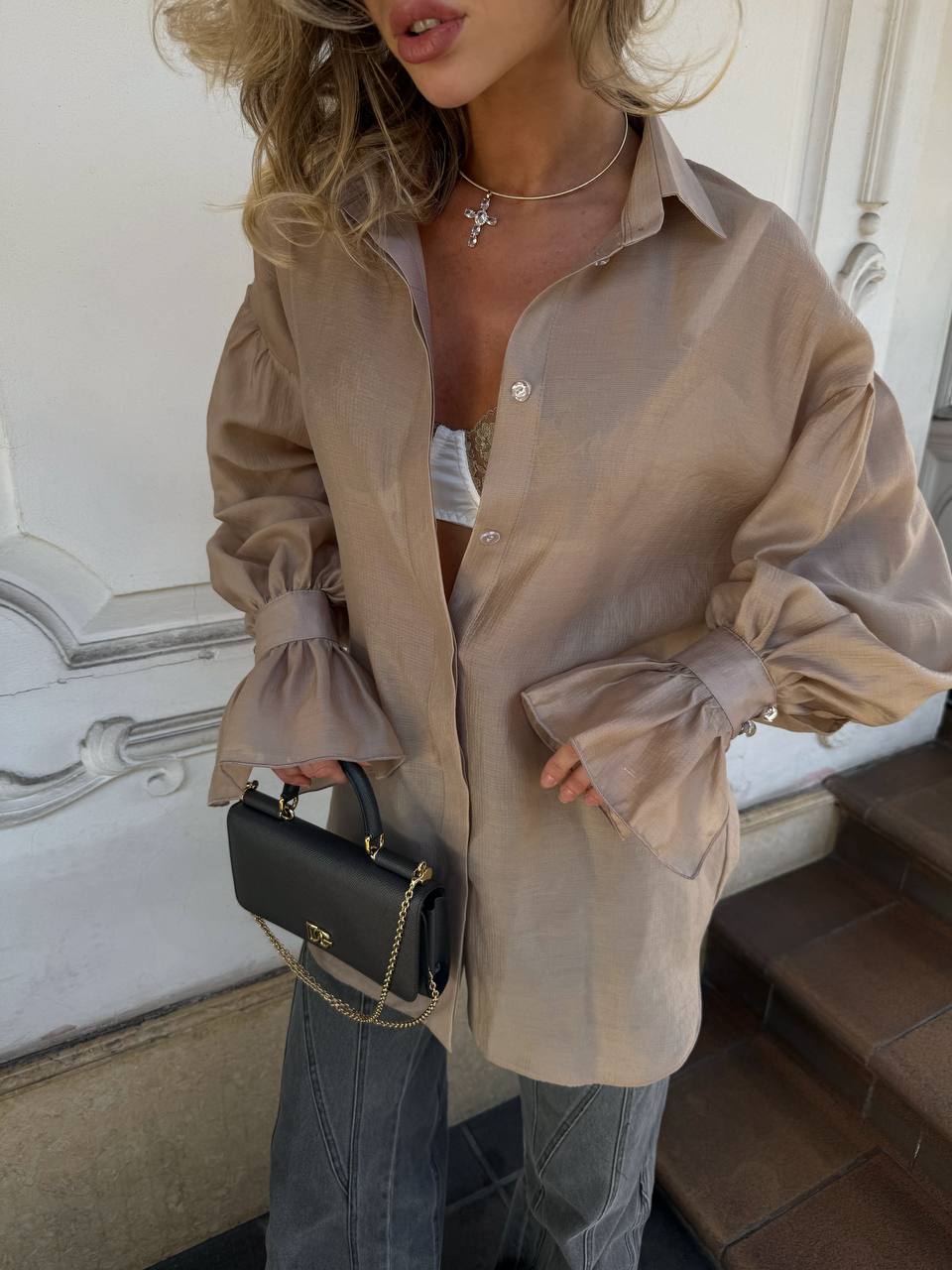 Woman wearing a beige sheer shirt with ruffled sleeves and holding a black handbag.