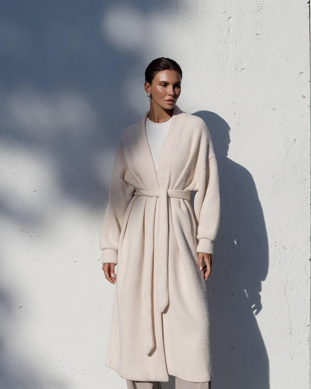 Woman wearing a long beige coat against a white wall