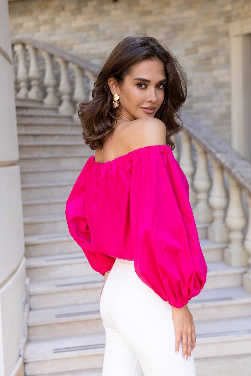 Woman wearing a bright pink off-shoulder top and white pants on stone steps.