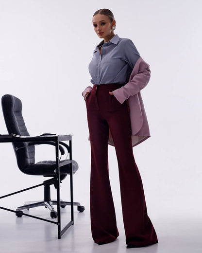 A woman models a stylish outfit consisting of a slate blue button-down shirt, a light pink cardigan draped off her shoulders, and high-waisted burgundy flare pants, standing next to a black office chair in a bright studio setting.