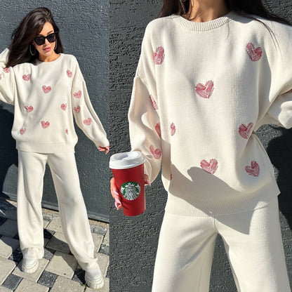 Woman wearing a white sweater with red heart patterns and white pants, holding a red Starbucks cup.