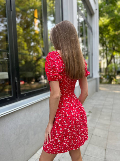 Woman wearing a red floral dress standing outdoors near a building with large windows.