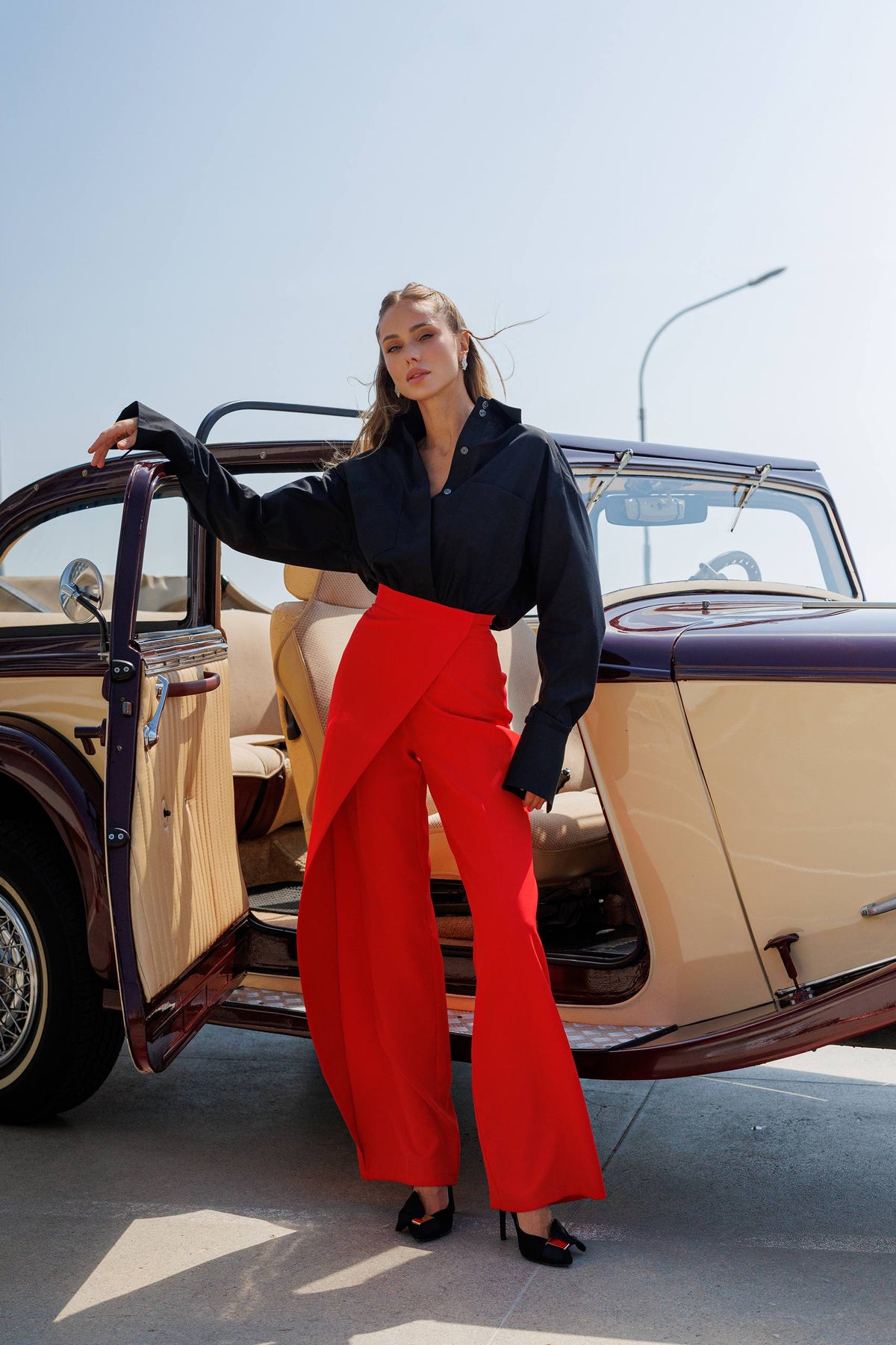 Woman in a black top and red pants standing next to an open car door.