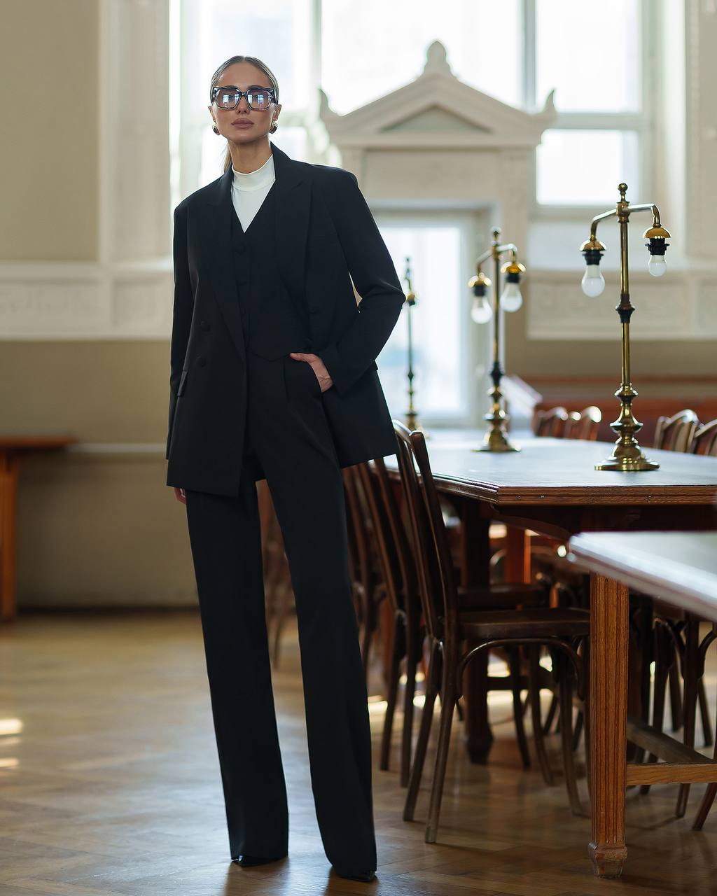 Person wearing a black suit standing in an elegant room with wooden furniture and large windows.