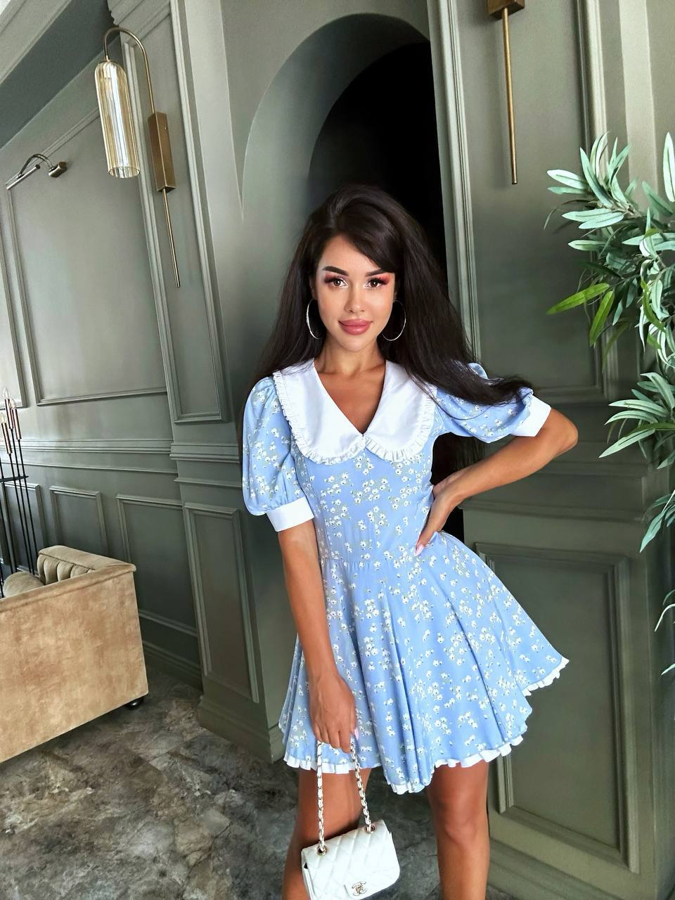 Woman in a blue floral dress standing in a room with green walls and a plant.