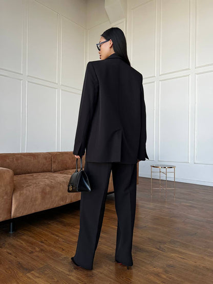 Person wearing a black suit standing in a room with a brown couch and white paneled walls.