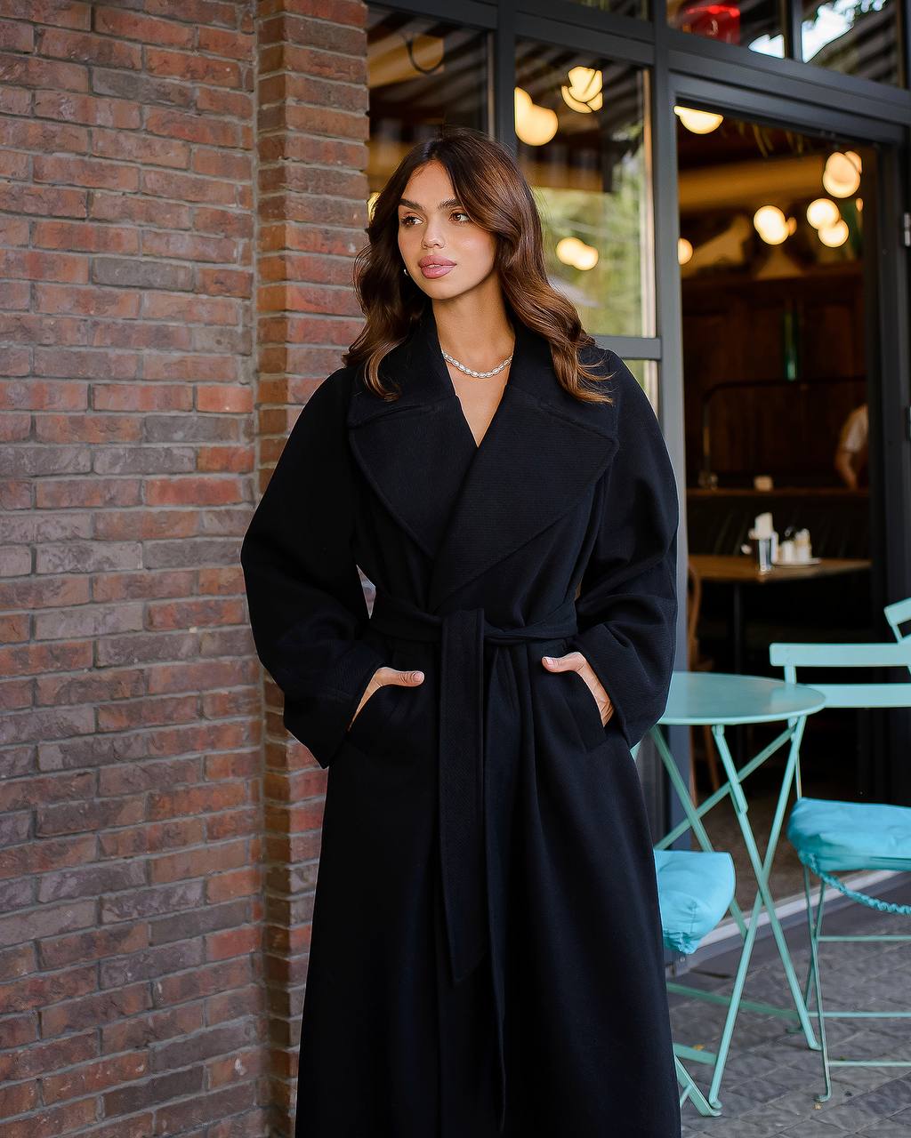 Woman wearing a black coat standing against a brick wall with outdoor cafe furniture in the background.