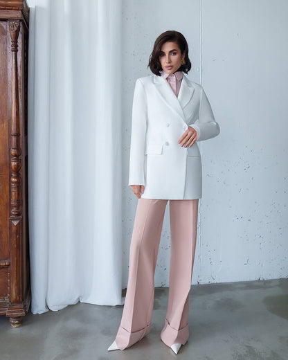 Woman wearing a white blazer and pink pants against a white wall.