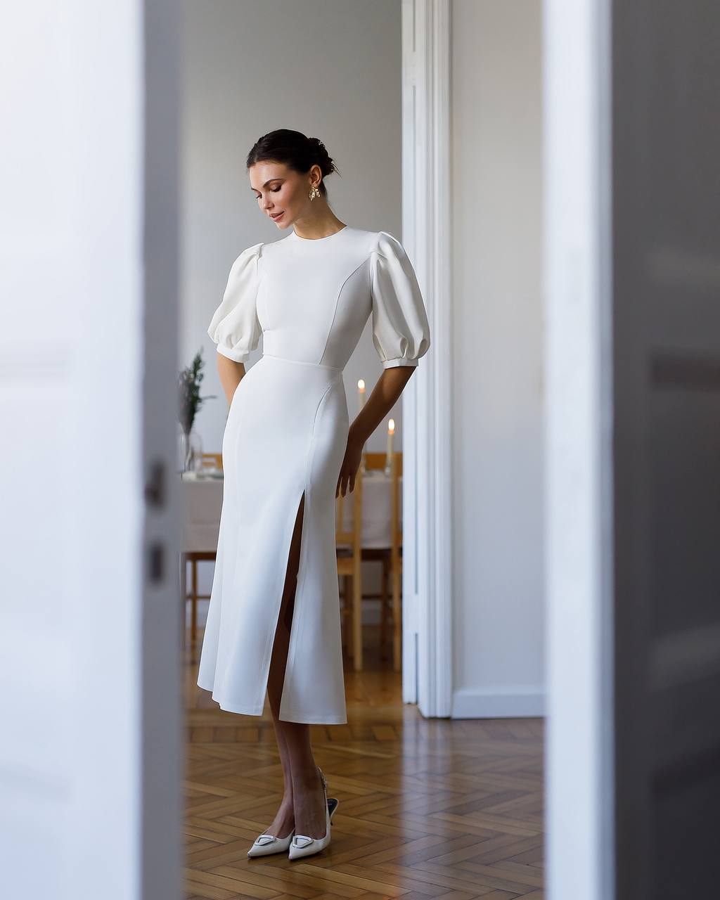 Woman in a white dress with puffed sleeves standing in a room with wooden flooring.