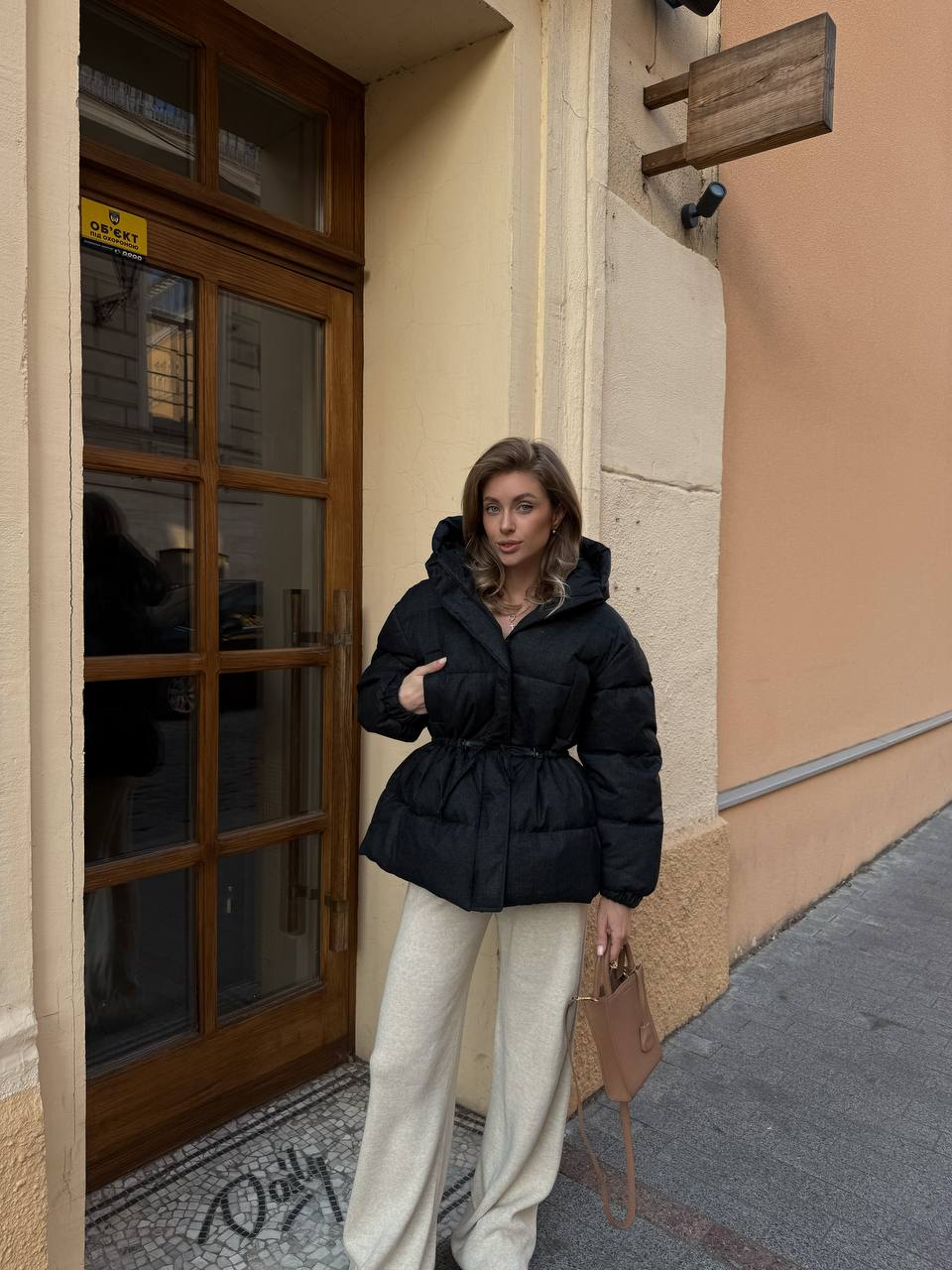 Woman in a black puffer jacket and beige pants standing on a street corner.