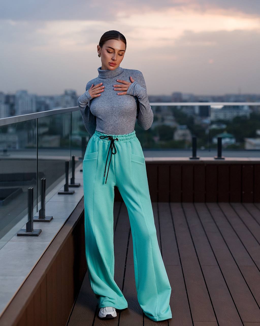 Woman in gray turtleneck and teal pants standing on a rooftop with cityscape background
