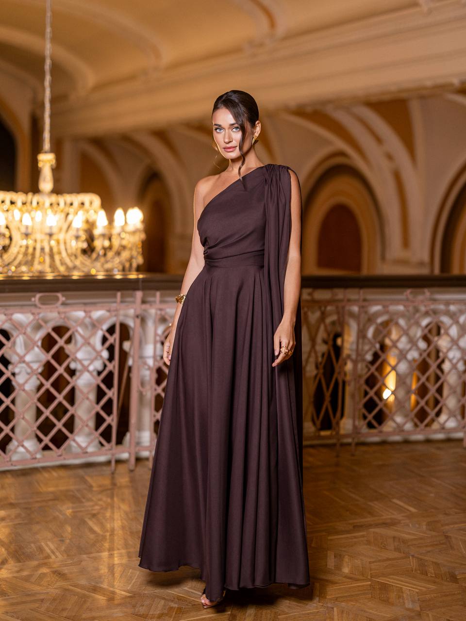 Woman in a dark purple one-shoulder dress standing in an elegant indoor setting with chandeliers.