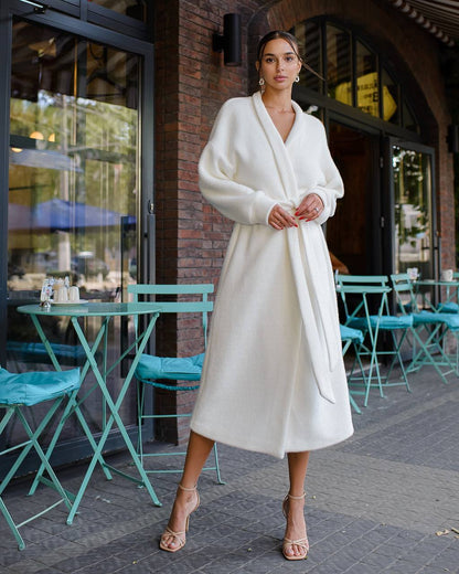 Woman in a white coat standing outside a building with teal chairs and tables.