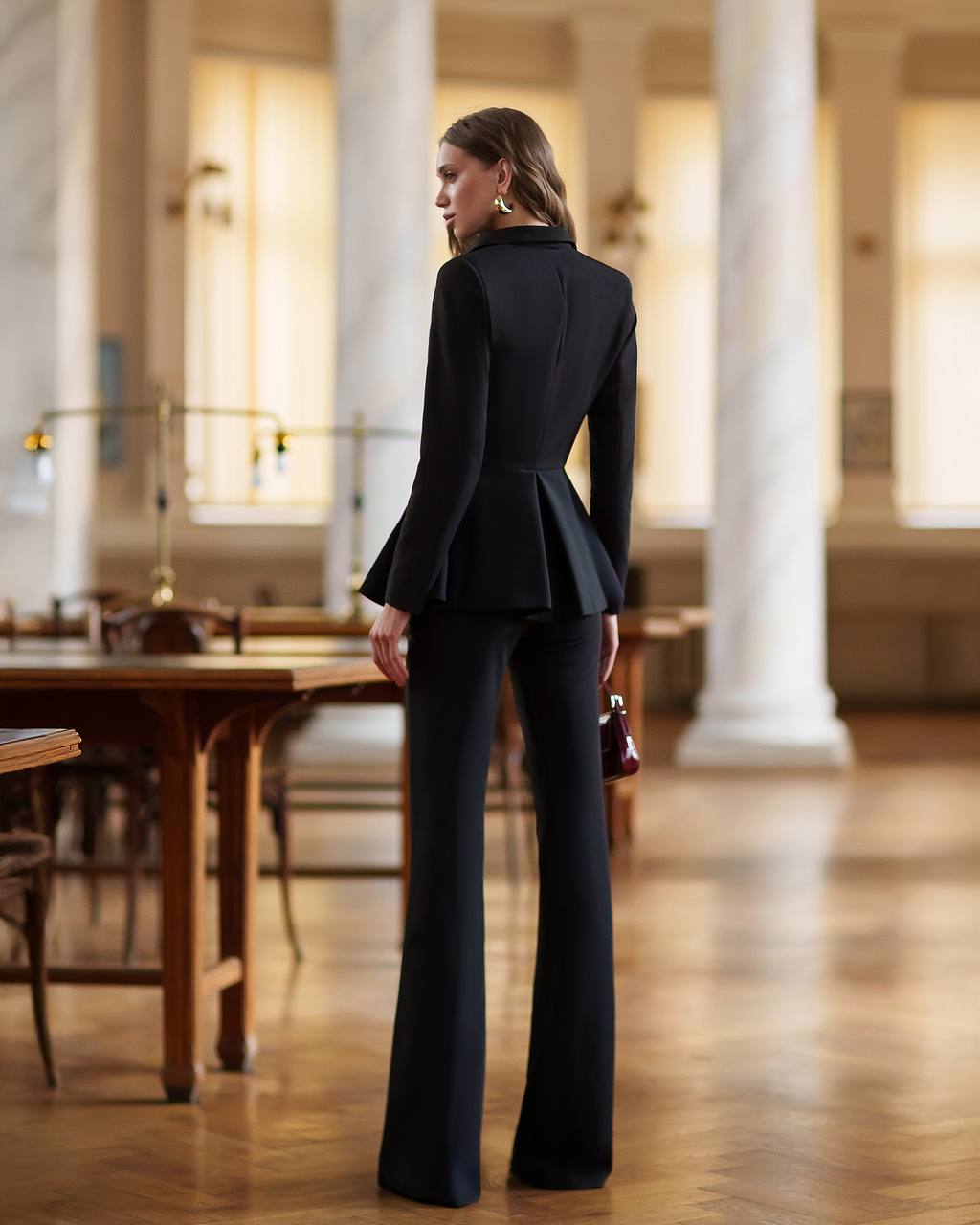 Woman in a black suit standing in an elegant indoor setting with columns and wooden tables.