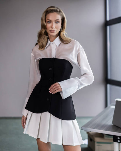 A woman models a unique two-tone outfit featuring a sheer white long-sleeve button-up shirt layered over a structured black corset-style vest, paired with a white pleated mini skirt, set against an office-like background.
