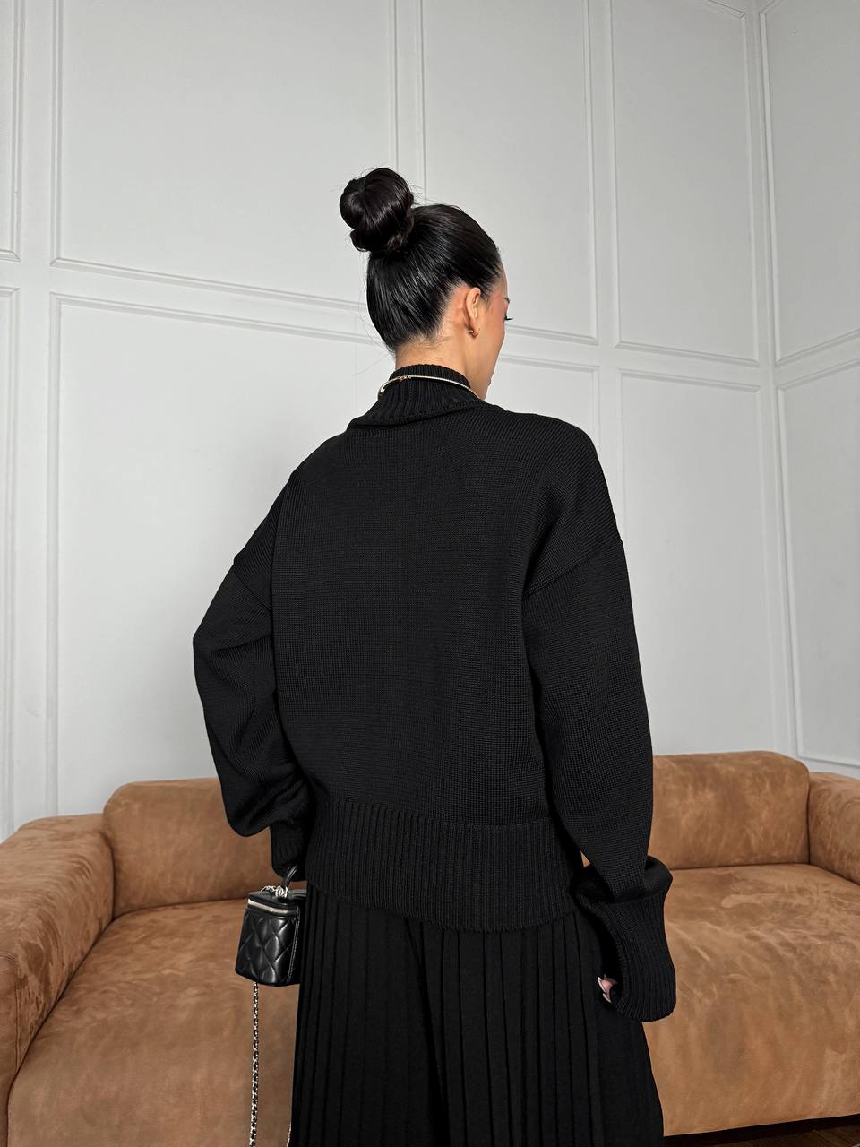 Person wearing a black sweater and skirt standing in front of a beige sofa against a white paneled wall.