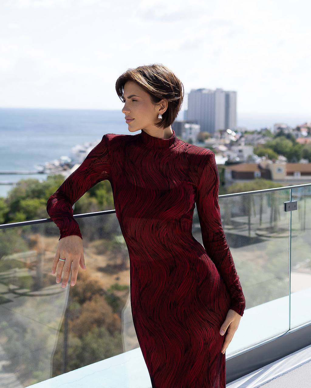 Woman in a burgundy dress standing on a balcony with a cityscape and water view.
