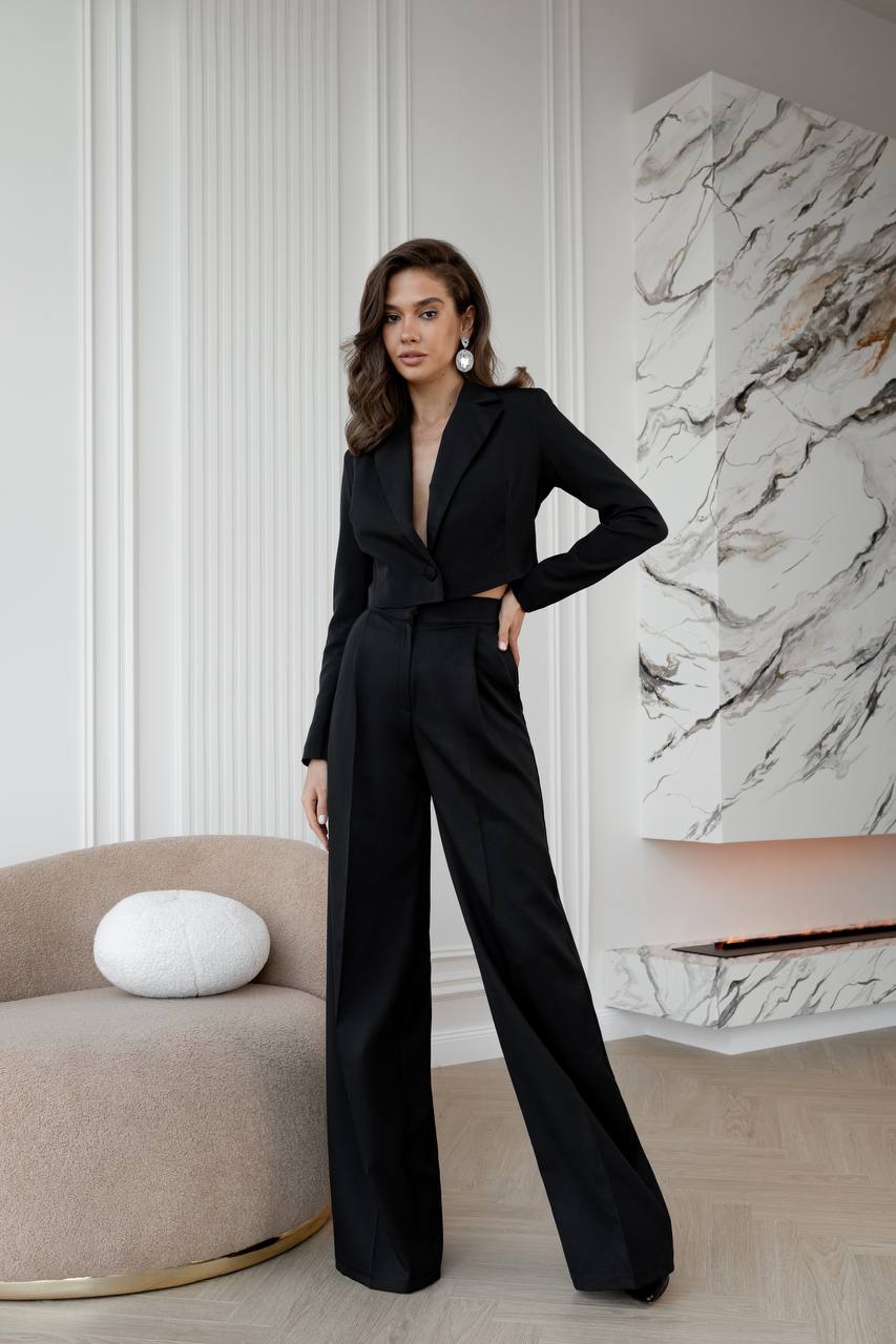 Woman in a black jumpsuit standing in a stylish room with marble wall and beige chair.