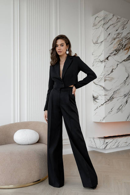 Woman in a black jumpsuit standing in a stylish room with marble wall and beige chair.