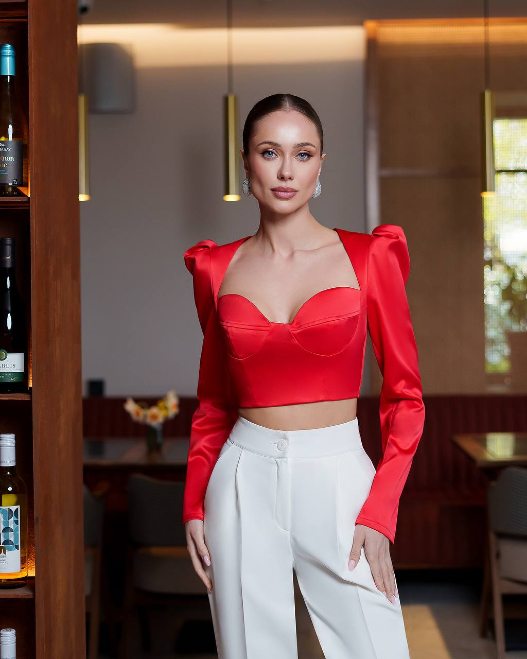 A woman stands in a restaurant setting wearing a bright red satin long-sleeve crop top with puffed shoulders and white high-waisted trousers.
